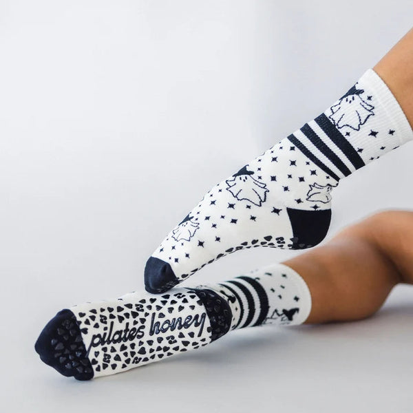 A pair of white crew socks with black patterns, including stripes, stars, hearts, and the text 'pilates honey', worn on a person's feet against a light gray studio background.