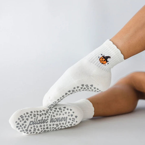 White crew socks worn on feet, with one sock showing a small orange pumpkin graphic and the other showing gray grip dots on the sole. The word 'pilates honey' is printed on the sole.