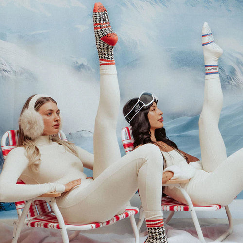 Two women in vintage-style winter outfits with checkered socks against a snowy mountain background.