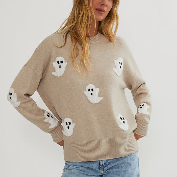A woman wearing a beige crewneck sweater with white ghost motifs knitted all over it, paired with light blue jeans, against a plain light gray background.