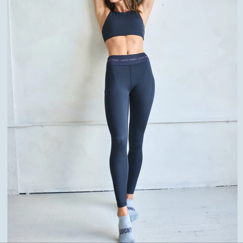 Woman wearing dark blue athletic leggings and sports bra against a light gray wall.