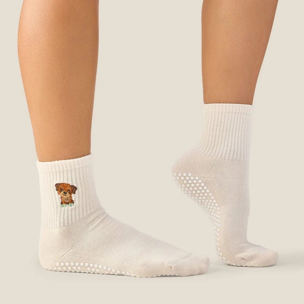 A pair of off-white ankle socks worn on a person's feet, featuring a small cartoon dog graphic on the left sock and grip dots on the soles, against a solid light beige background.