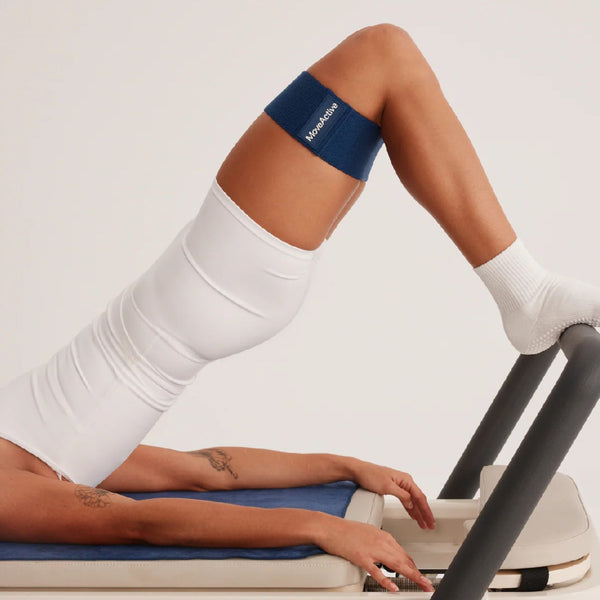 Person using a reformer with a blue band on their knee, wearing white socks and pants.
