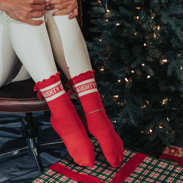 Person wearing red 'NIGHTY' socks sitting on a wrapped Christmas present with a decorated tree in the background.