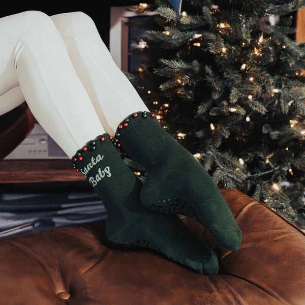 Person wearing green socks with 'Santa Baby' text in front of a decorated Christmas tree.