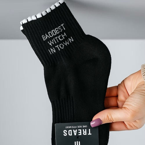 Black socks with 'Baddest Witch in Town' text held by a hand against a gray background
