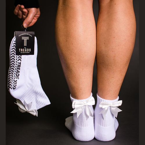 White socks with bows worn by a person, held by a hand with a Treads branded sock on a black background.