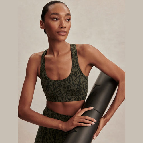 Woman holding a black yoga mat wearing a green camouflage sports bra and leggings on a beige background